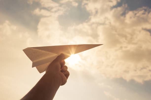 A hand launching a paper airplane into a sunlit, cloudy sky